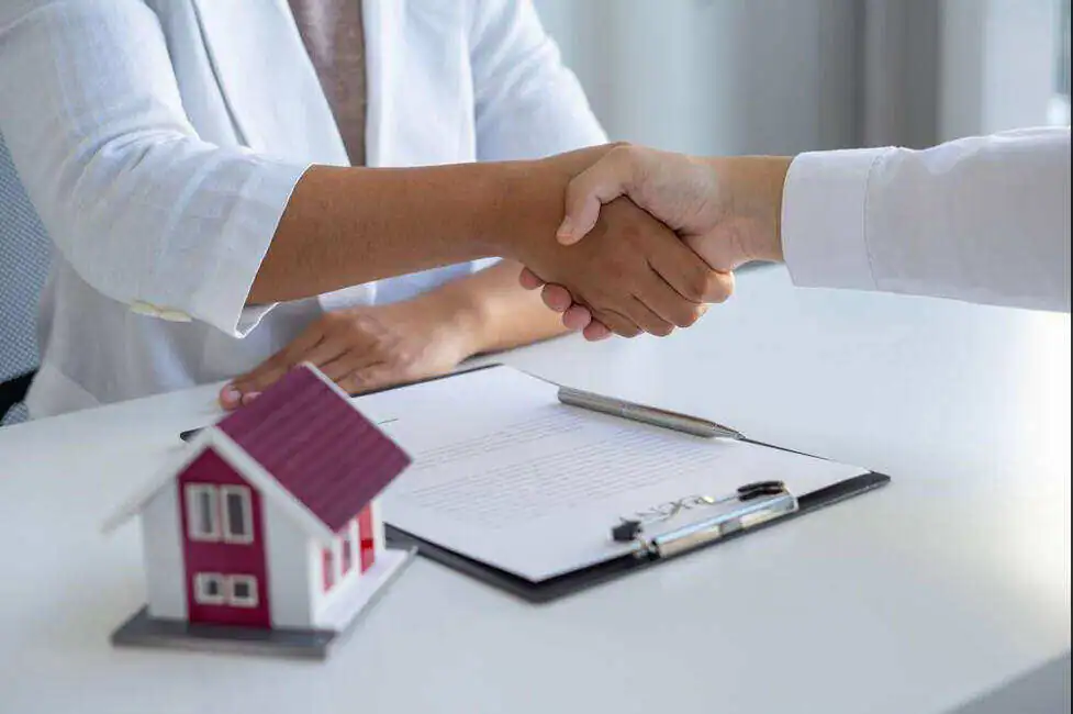 two people shaking hands implying a transaction with a clipboard and house model 