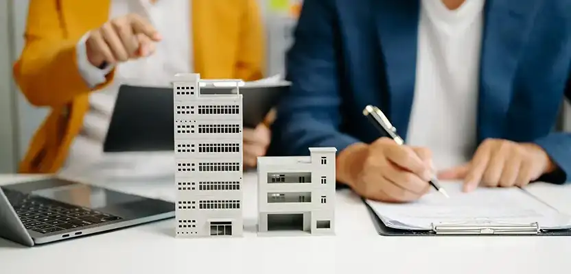 2 people sitting infront of a table. One person writing on a clipboard, one person pointing to a model of a building on the table 